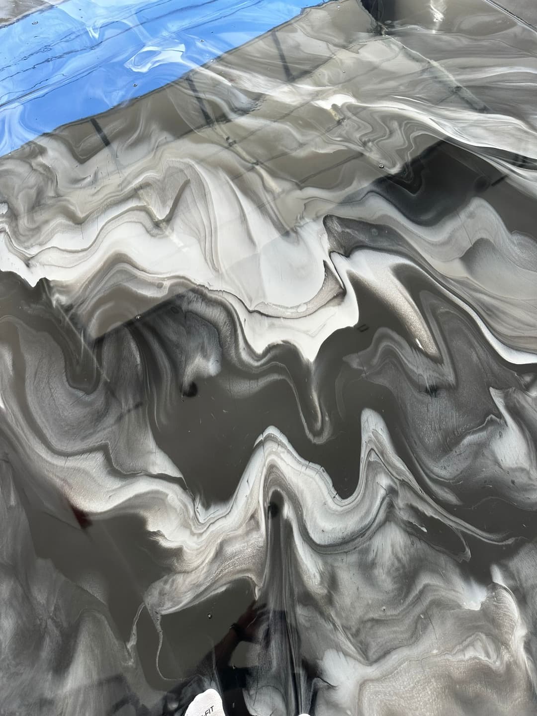 Swirling black and white abstract resin art with hints of blue tape on the edge.