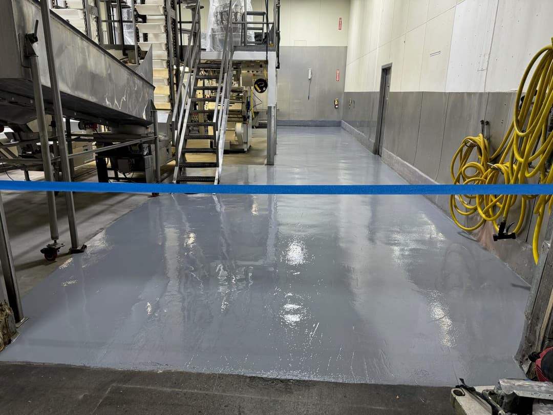 Freshly coated gray epoxy floor in industrial setting with safety tape and equipment in background.